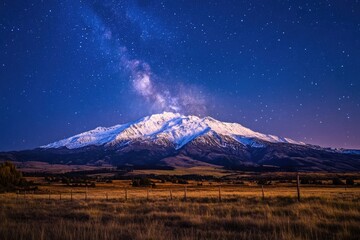 Fototapeta premium Snow-capped mountain under a breathtaking milky way, illuminated field in the foreground, stars twinkling in the dark blue night sky.
