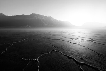 Black and white shot of a cracked earth surface with mountains in the background under a bright sky.
