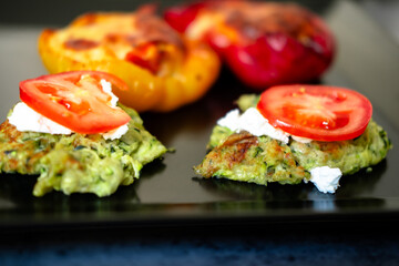 Grilled bell peppers and zucchini fritters with tomato topping on black plate.