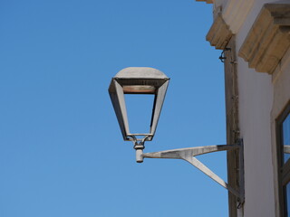 The image shows a street lamp or wall lamp attached to a building wall. The lamp has a classic, conical design and is made of metal. It is depicted against a clear blue sky.