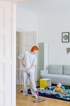 A woman mops the floor in a living room, a child is lying on carpet