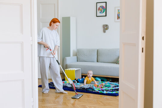 A woman mops the floor in a living room, a child is lying on carpet