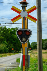 A railroad crossing signal stands tall next to a rural road, featuring a unique triangular design with distinctive red lights. Clear skies and greenery surround this safety feature