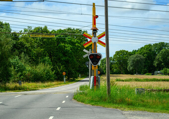 Red signals illuminate at a railroad crossing where a rural road meets train tracks, surrounded by green trees and open fields under a partly cloudy sky
