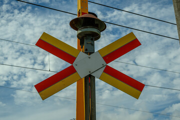 A bright railroad crossing signal featuring red and yellow colors stands prominently against a blue sky with scattered clouds. Nearby, train tracks are visible, signaling caution