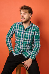 Young Caucasian man in his twenties sitting on stool with arms crossed looking thoughtfully to side...