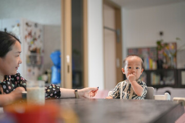 Chinese family shares a meal in the living room, a mother in her thirties and her one-year-old baby at the table, baby in a high chair eating bread amid furniture and light. Spring. Shanghai. China.