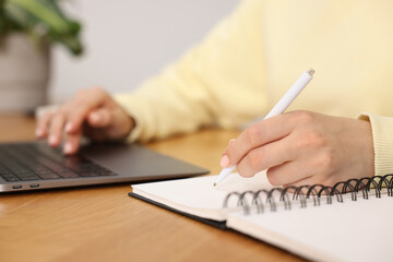 Fototapeta premium Copywriter using laptop and writing in notebook at wooden table indoors, closeup