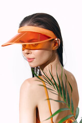 Fototapeta premium Young woman with an orange visor and tropical leaves, expressing confidence and style against a minimalistic background.