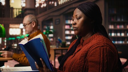 African american woman reading book and completing her class notes in a study room, preparing for her thesis and researching archived academic materials at the public library.
