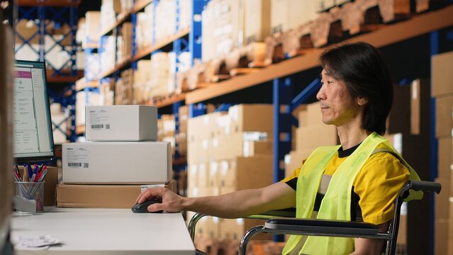 Asian man wheelchair user typing tracking numbers on a computer software, updating parcel status in real time. Staff managing order fulfillment in a logistics center to sort inventory. Camera B.