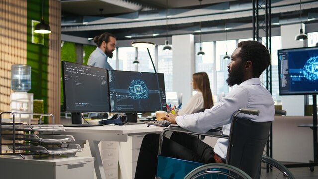 IT professional in wheelchair in inclusive office developing software applications using AI automation. Computer scientist with paraplegia debugging applications with artificial intelligence, camera A