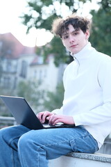 male student with coffee to go sitting in park and using laptop
