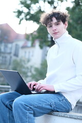 male student with coffee to go sitting in park and using laptop