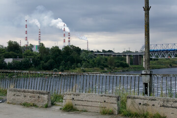 Industrial scene with tall chimneys emitting smoke beside a river and bridge, framed by green...