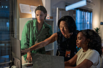 Three women collaborate on a computer in an office during late night