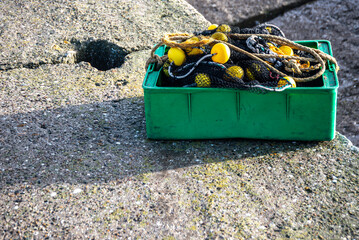 Green plastic box filled with fishing nets and yellow buoys placed on a concrete pier by the sea. Representative image of artisanal and commercial fishing activity.