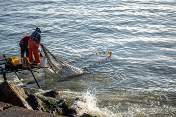 Two fishermen handling a net with yellow buoys on the seashore, illustrating artisanal and traditional fishing activity.