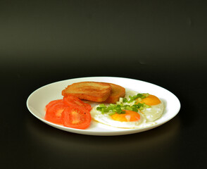 Two fried eggs scorched with cut green onions with pieces of tomato and two toasts of croutons in a flat and wide plate on a black background.