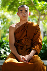 A young buddhist monk sits quietly in a serene garden, dressed in an orange robe, surrounded by vibrant greenery and sunlight. Harmony and Religion concept. Generative AI.