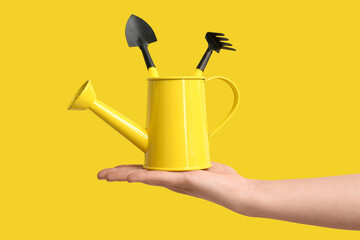 Female hands holding watering can with gardening rake and shovel on yellow background