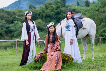 Three women in medieval dresses pose outdoors with white horse, surrounded by lush greenery and mountains