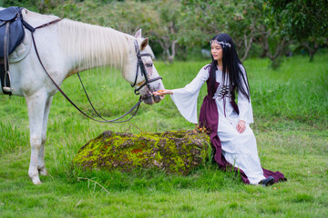 Woman in medieval dress sits on grass feeding white horse in lush green field