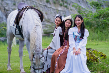Three women in elegant dresses sit on grass beside white horse in serene outdoor setting. scene exudes sense of tranquility and grace