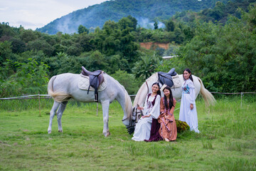 Three women in traditional dresses pose with two white horses in lush, green landscape with mountains in background
