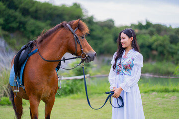 Woman in white dress stands beside brown horse in lush green field, holding its reins and smiling gently