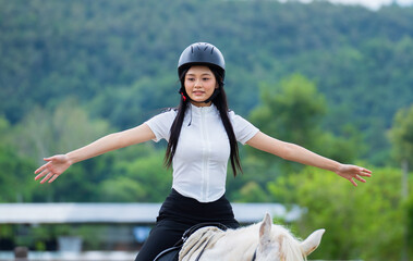 Woman in white shirt and helmet joyfully balances on white horse with arms outstretched, set against lush green landscape