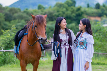 Two women in medieval style dresses stand beside brown horse in lush, green landscape, smiling at each other