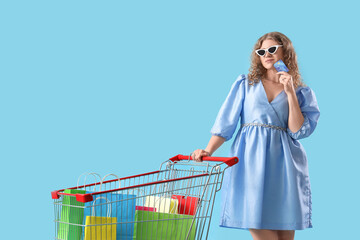 Beautiful young stylish thoughtful woman with credit card and shopping cart full of bags on blue...