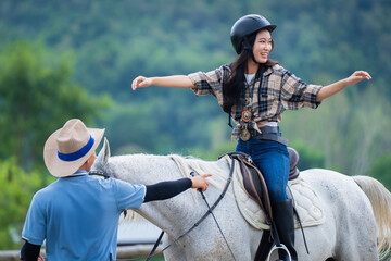 Woman joyfully balances on white horse while guide assists, set against lush green background