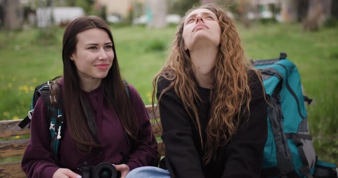 Two young women reviewing travel photographs on their camera, reliving happy memories while sitting together on a park bench.