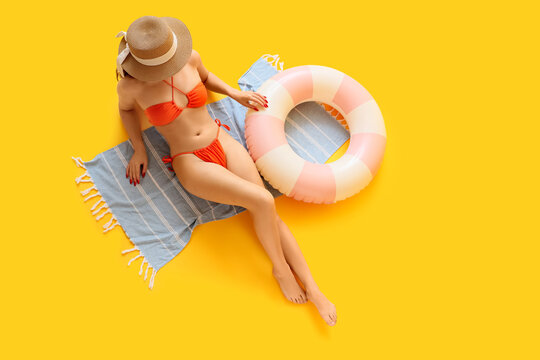 Beautiful young woman in stylish orange swimsuit with inflatable ring and beach blanket lying on yellow background, top view