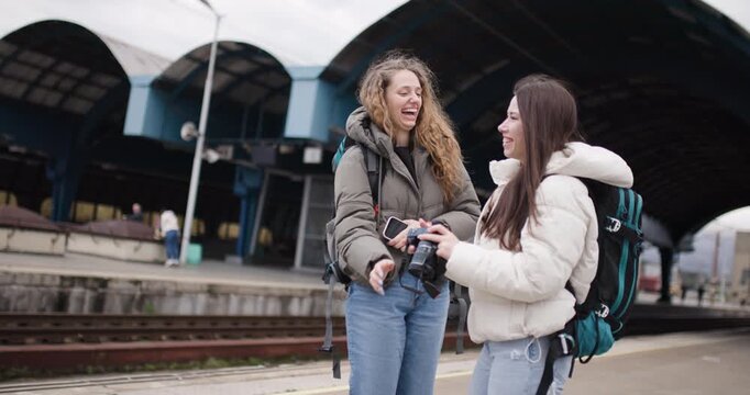 Two Young Women With Backpacks at a Train Station, One Taking Photos of the Other, Excited About Their Travel Adventure