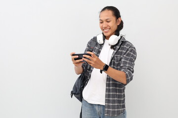 Teenage college boy in casual clothes, backpack, headphones around neck, playing game on mobile smartphone, standing isolated on white background.