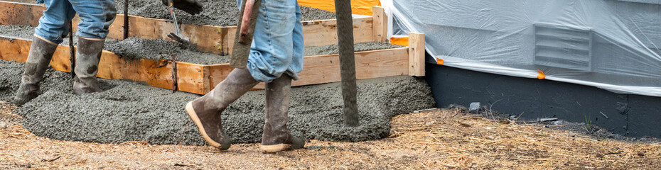 Construction workers in rubber boots and hard hats working cement pumper hose to pour fresh cement into wood forms for concrete back patio and stairs, and smooth it out, new housing development
