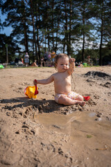 adorable baby enjoying playful day in the sun at muddy beachfront with toy bucket and pine forest backdrop, surrounded by nature in a happy summer setting, relax, carefree, summerday, play, leisure