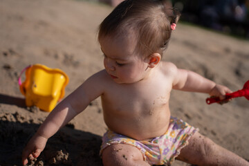 young child plays in sunlit sandy beach, exploring tactile sand textures with curiosity, colorful bucket and red shovel beside, illustrating joyful childhood innocence, backlight, portrait