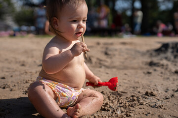 a young child in colorful shorts sits on sandy ground, playing with a stick and red shovel under bright sunlight, surrounded by blurred outdoor greenery and people in the background, casual, bokeh