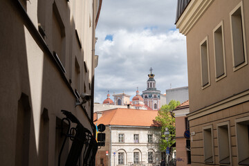 narrow urban alleyway with ornate historic church towers under a partly cloudy sky, framed by residential buildings, displaying a juxtaposition of old and modern architecture, sunlit, serene