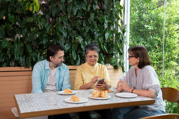 Group of diverse senior and middle-aged friends sitting together in a cafe, chatting and looking at a smartphone while enjoying pastries, creating a warm and friendly social atmosphere.