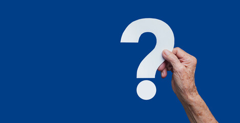 Elderly hand holding paper question mark on blue background for curiosity and inquiry concept.