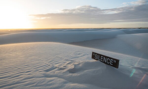 Sandy dunes under a bright sky, sign reads "SILENCE". Light flare