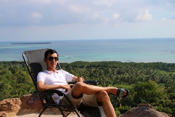 Man in sunglasses relaxing on a lounge chair with a panoramic view of lush tropical coastline,...