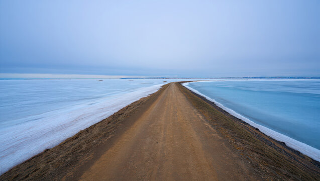 A long dirt road leads through a frozen landscape with ice on either side under a vast, overcast sky, suggesting a journey