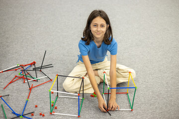 Girl in a blue T-shirt sitting on the floor assembling a colorful plastic stick and connector...