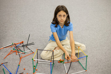 Girl in a blue T-shirt sitting on the floor assembling a colorful plastic stick and connector...